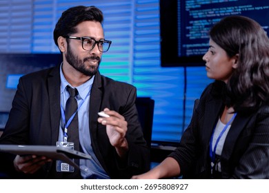 Code deep dive briefing. Experienced male developer explains software architecture or complex algorithm to female engineer with code visible on screen in background tech hub.
