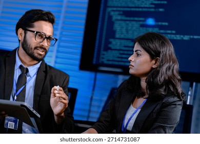 Code deep dive briefing. Experienced male developer explains software architecture or complex algorithm to female engineer with code visible on screen in background tech hub.