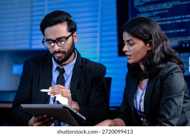 Code deep dive briefing. Experienced male developer explains software architecture or complex algorithm to female engineer with code visible on screen in background tech hub.
