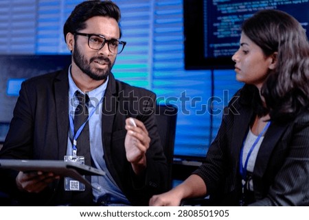 Code deep dive briefing. Experienced male developer explains software architecture or complex algorithm to female engineer with code visible on screen in background tech hub.