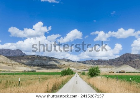 Cody, Wyoming, USA - View across rural landscape with country road flanked by grasses, fields, and with foothills of Rocky mountains under blue sky in Cody, Wyoming, USA.
