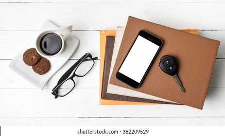 coffe cup with cookie,phone,car key,eyeglasses and stack of book on white wood table