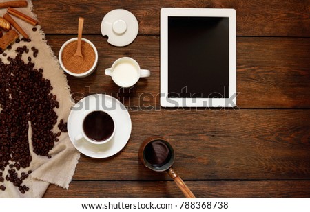 Coffee background. White tablet computer blank black screen with white cup of coffee and coffee beans on dark wooden background. Freshly made coffee in old rustic desk. Top view with copy space. 