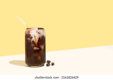 Coffee beans and cold coffee drink with glass straw on a yellow isometric diagonal projection background. Can-shaped glass cup of coffee with plant-based milk. Concept of sustainable summer drinks. - Shutterstock ID 2161826429