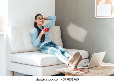 Coffee Break. Beautiful Cheerful Young Woman In Glasses Holding Coffee Cup And Looking Relaxed While Sitting On The Couch In Office