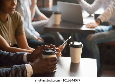 Coffee break in cafe with gadgets concept, diverse young african and caucasian people sitting at coffeehouse tables with drink to go in paper cups enjoy using mobile phone and laptop, close up view