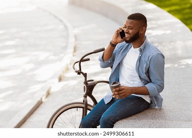 Coffee Break. Cheerful African Guy Relaxing With Takeout Drink On Bench In Park And Talking On Cellphone With His Bike Parked Next By - Powered by Shutterstock