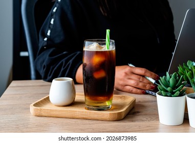 Coffee Break Concept. An Entrepreneur Woman Relaxing Drinking Iced Coffee In Between Work Online On Laptop. Close Up A Glass Of Cold Black Americano Coffee On The Desk Woman Working In The Background.