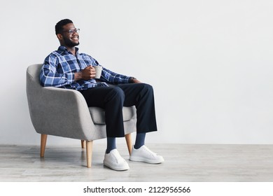 Coffee Break. Dreamy Young African American Man Sitting In Armchair, Enjoying Cup Of Hot Beverage Against White Studio Wall, Copy Space. Millennial Black Guy Relaxing With Mug Of Warm Drink