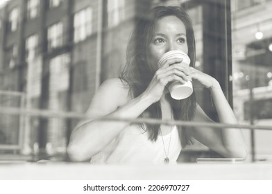 Coffee break inside a shop. Beautiful asian woman drinking a coffee cup in a store. View through the window screen
