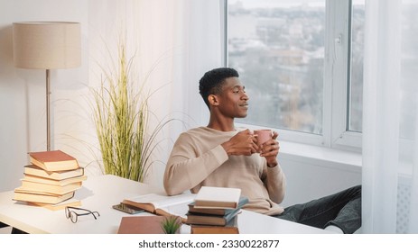 Coffee break. Morning ritual. Study relax. Dreamy peaceful young man drinking cup of hot tea enjoying leisure home window view at desk with books. - Powered by Shutterstock