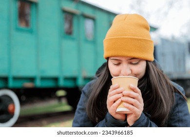 Coffee Break in the Rail Yard: Young Latina Woman in Warm Attire Savoring a Takeaway Coffee Cup. Copy space