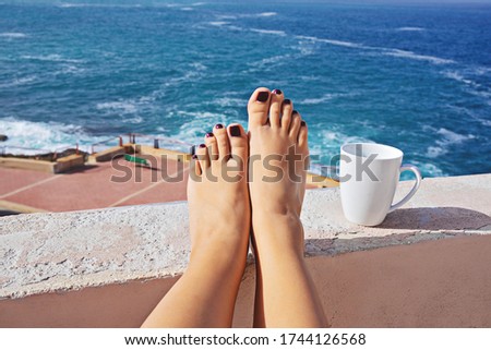 Coffee break and self-isolation concept. Female barefoot against blue sea and white mug of coffee or tea, POV view
