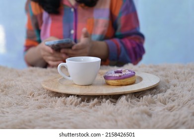 A coffee break set consisting of coffee in a white cup and purple donuts served in a wooden tray is placed on cream colored carpet. Woman checking important information from mobile phone during break.