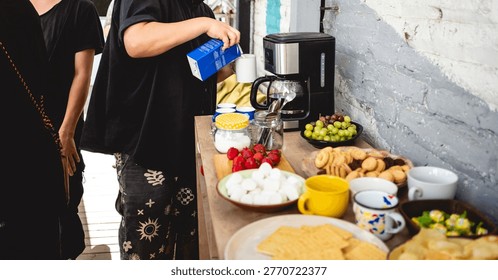 Coffee break table with snacks and drinks (chips, strawberries, grapes, cookies, marshmallows, candies, cake), with young person preparing coffee with milk and sugar