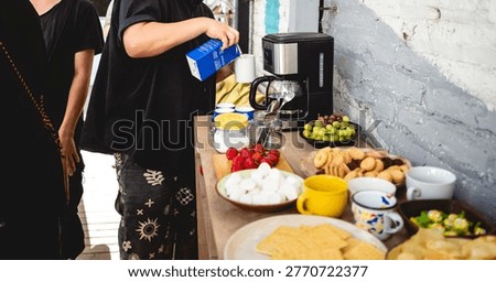 Coffee break table with snacks and drinks (chips, strawberries, grapes, cookies, marshmallows, candies, cake), with young person preparing coffee with milk and sugar