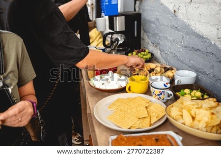 Coffee break table with snacks and drinks (chips, strawberries, grapes, cookies, marshmallows, candies, cake), with young person preparing coffee with milk and sugar