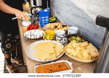 Coffee break table with snacks and drinks (chips, strawberries, grapes, cookies, marshmallows, candies, cake), with young person preparing coffee with milk and sugar