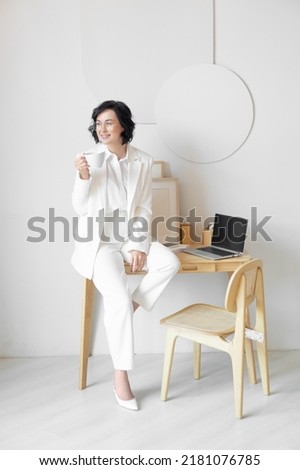 Coffee break. A woman in a white suit with a beautiful smile drinks coffee while sitting on a wooden table. Wooden furniture. Bright modern workplace.