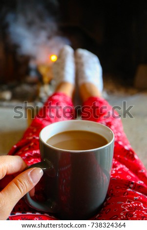Coffee cap, firewood fire on the background. Bright colorful flame, burning wood at the fireplace, female legs in socks warming up. 