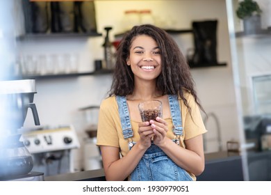 Coffee Connoisseur. Happy Woman With Long Dark Hair In Casual Clothes With Portion Of Coffee Beans In Glass In Cafe