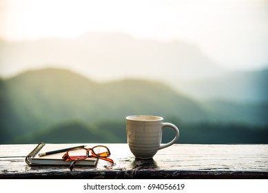 A Coffee Cup, Eyeglasses And A Note Book On Wooden Table With Sunrise And Mountain  Bokeh Background. A Start Of New Day With Hot Beverage. Business Concept.