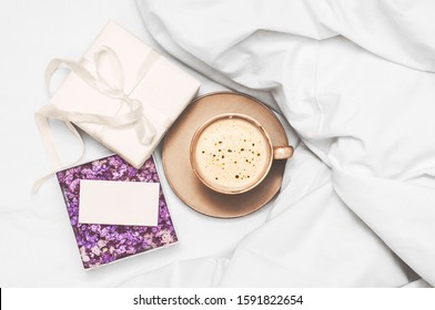 Coffee cup with foam, gift box with spring flowers on a white bedsheet and coverlet.  Coffee with a surprise in bed.  Top view.  Flat lay - Powered by Shutterstock