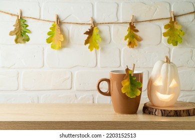 Coffee Cup And Jack O Lantern Pumpkin Decoration On Wooden Shelf Over Brick Wall Background With Autumn Leaf Garland. Halloween Holiday Greeting Card