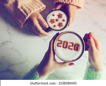 Coffee Cup With The Number 2020 On Frothy Surface In Female Hands Holding Over Blurred Marble Table Background And Another One With Star Symbols On Frothy Surface. Happy New Year 2020 Food Art Theme.