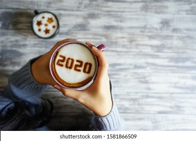 Coffee Cup With Number 2020 On Frothy Surface In Female Hands Holding Over Grey Painted Wood Plank Background And Another Cup With Star Symbols On Frothy Surface. Happy New Year 2020 Food Art Theme.