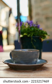 Coffee Cup On Wooden Table Outdoors With Blurred Out Plant Pot In Background