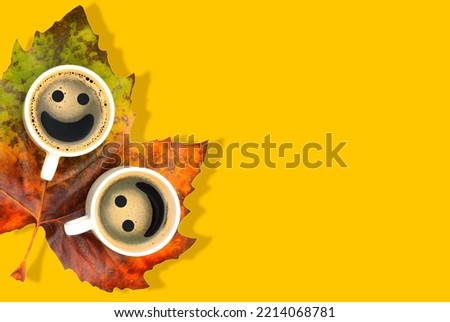 Coffee cups with smiley faces placed on a large pigmented maple leaf isolated on yellow with copy space. Cosy autumn background.