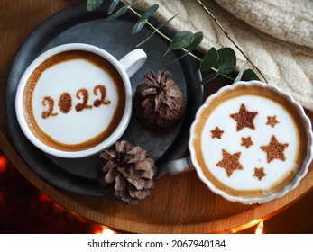 Coffee cups with star symbols and number 2022 on frothy surface flat lay on round wooden table with eucalyptus branch, pine cones, knitted sweater. Holidays food art for Christmas and Happy New Year.