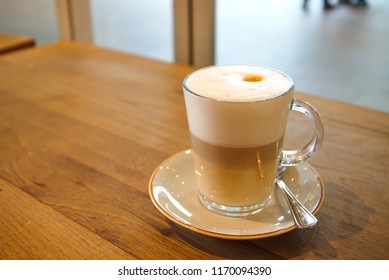 Coffee Latte Macchiato In Clear Transparent Glass And White Plate Lay On Rough Wooden Table Counter Beside Windows Of Coffee Sop And Blur Background Outside Coffee Shop.