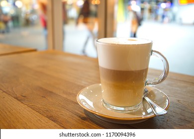 Coffee Latte Macchiato In Clear Transparent Glass And White Plate Lay On Rough Wooden Table Counter Beside Windows Of Coffee Sop And Blur Background Outside Coffee Shop.