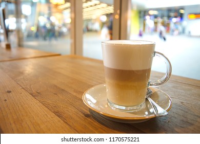 Coffee Latte Macchiato In Clear Transparent Glass And White Plate Lay On Rough Wooden Table Counter Beside Windows Of Coffee Sop And Blur Background Outside Coffee Shop.