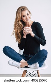 Coffee lover. Beautiful young woman holding coffee cup and looking away with smile while sitting on chair in lotus position against white background