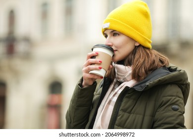 Coffee lover concept: young woman drinking hot beverage at street of European city. Model wearing green coat, white and beige scarf, yellow beanie. Close up. Outdoor shot