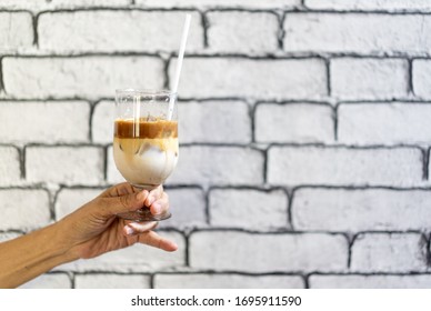 Coffee Macchiato And Milk Layer With Ice In Champagne Glass Was Held By Asian Grandmother Hand In Front Of White Brick Wall Wallpaper.
