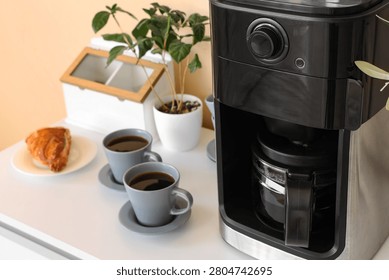 Coffee maker with two cups of black coffee and pastry on table
