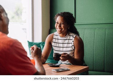 Coffee meet up - A cheerful black woman enjoys a lively conversation, smiling and having fun over coffee in a cozy cafe setting. She smiles at her partner affectionately. - Powered by Shutterstock