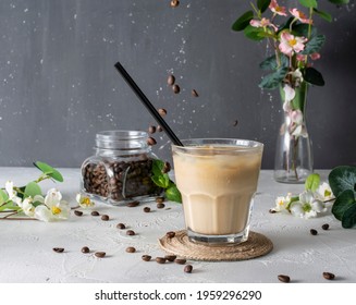 Coffee With Milk And Ice Is On The Light Table. Coffee Beans Pour From Above, A Bouquet In The Background