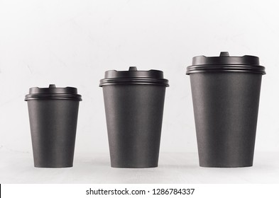 Coffee mockup - set of three different size blank black paper cups with cap on white wood table, coffee shop interior. Modern elegant concept for branding identity, advertising, design.
