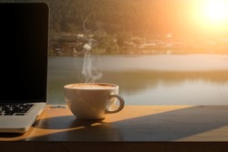 Coffee Morning And Laptop On Wooden Table With Lake, Mountain And Village Background.