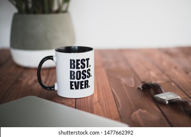 A coffee mug with best boss ever written on it on a wooden desk office with plant, laptop and watch. 