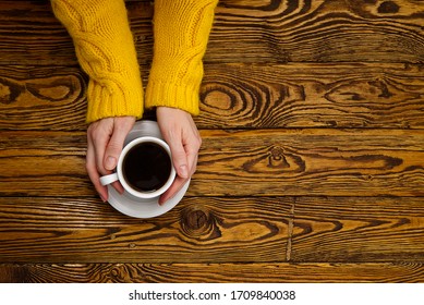 Coffee Mug In Hand On Old Wooden Table Top View With Copy Space. Woman In Cozy Yellow Home Sweater Holding A Mug Of Coffee While Enjoying A Drink