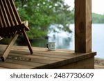 A coffee mug with the phrase "Life is better at the Cottage" rests beside a chair on the wooden porch of a boathouse, overlooking the serene blue waters of a lake in Muskoka, Ontario, Canada.