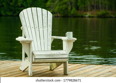 A Coffee Mug Sitting On A White Adirondack Chair On A Wooden Dock Near The Water.