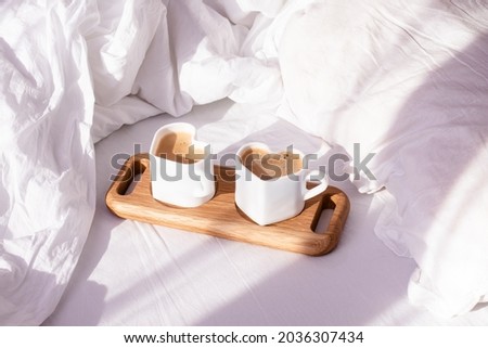 Coffee in mugs in the shape of a heart on a tray lie on a white bed. In the morning, the sun's rays illuminate the cups and a crumpled white sheet.