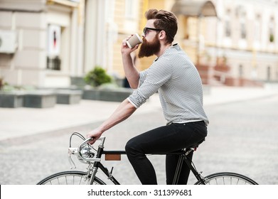 Coffee on the go. Side view of young bearded man drinking coffee while sitting on his bicycle outdoors  
